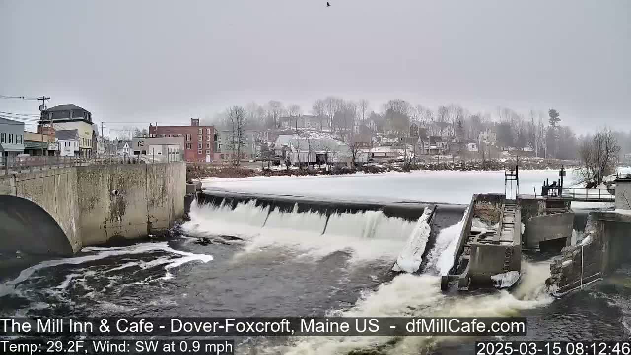 A partially frozen river flows over a dam in a small town on an overcast day.