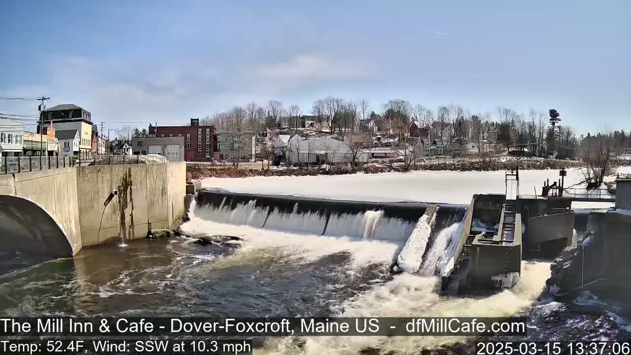 A dam with water flowing over it, a partially frozen river, and a small town in the background under a partly cloudy sky.