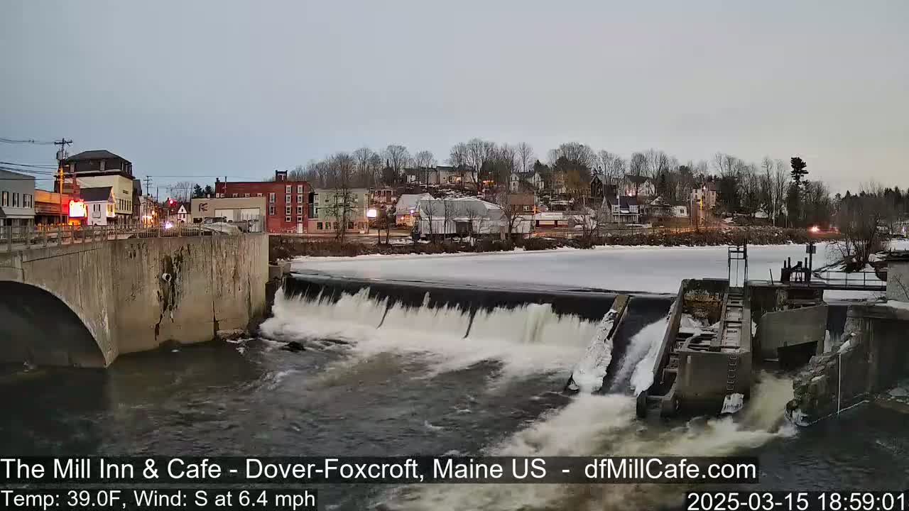 A river flows over a dam in a town under a twilight sky, with some ice visible on the river.