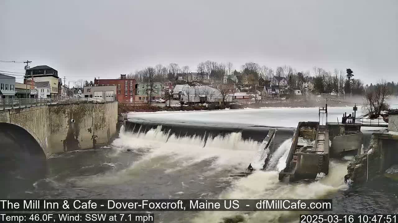 Water rushes over a dam in a town, with a partly frozen river and overcast skies.