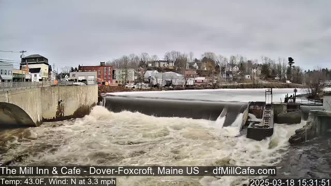 A rushing river flows over a dam in front of a small town on an overcast day.