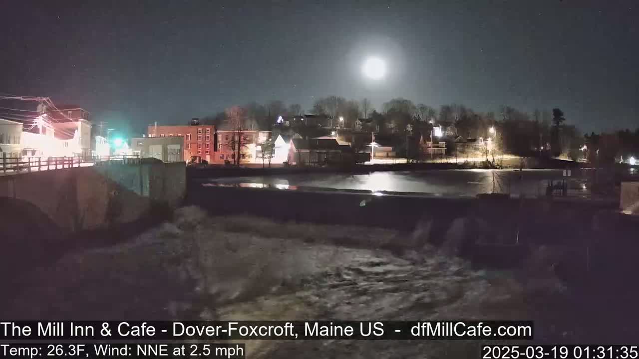 A nighttime view of a town's riverfront, with a dam showing water flowing over it under a bright moon.
