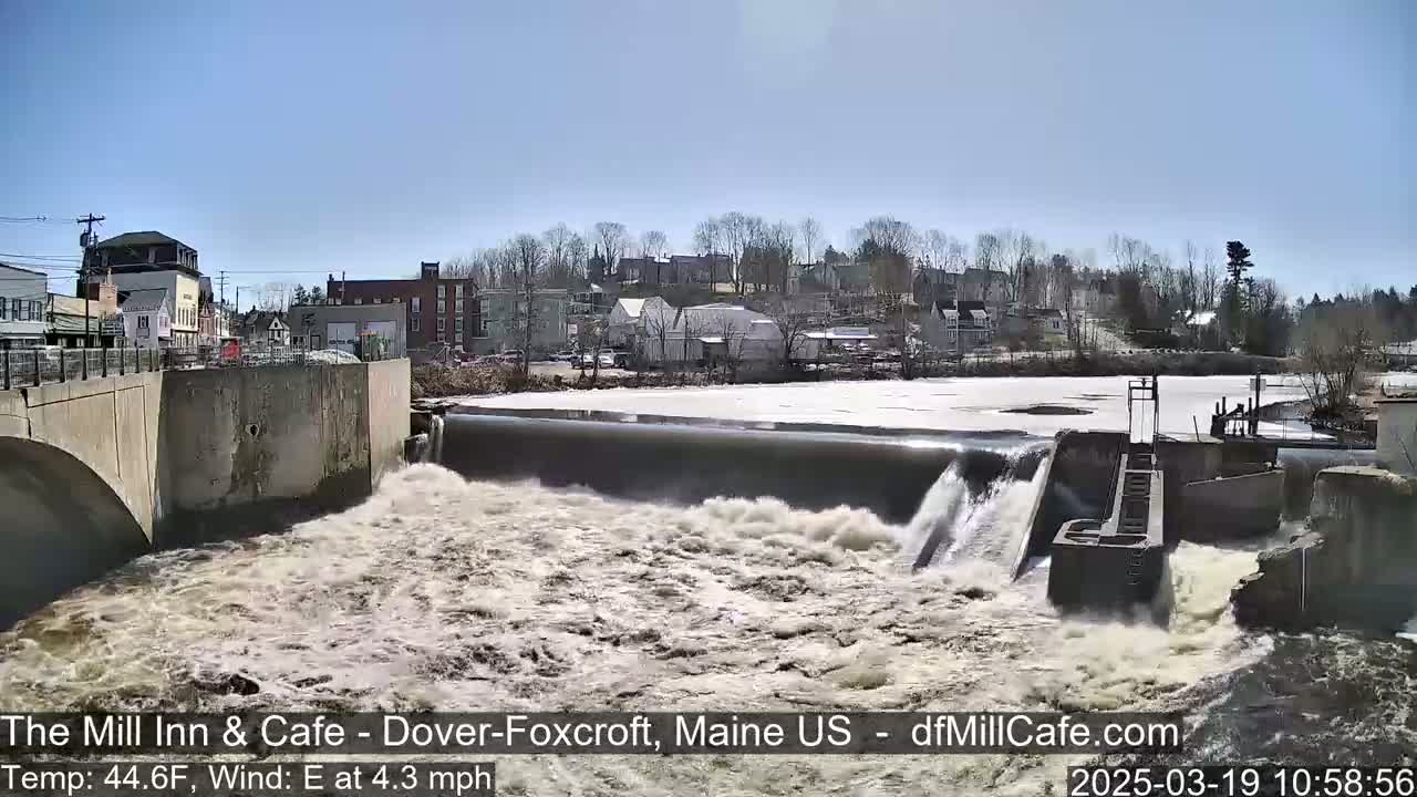 A dam releases water into a rushing river, with a town visible in the background under a clear blue sky.