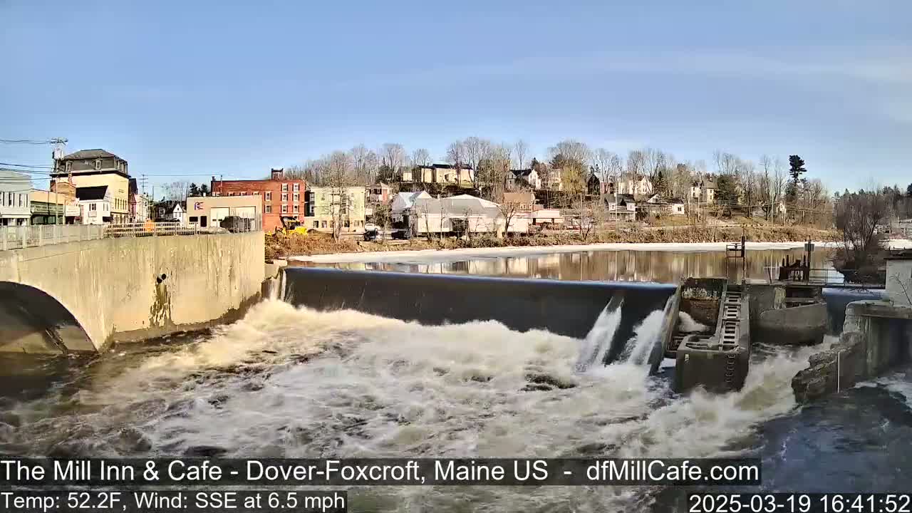Water rushes over a dam in front of a town on a sunny day with a light wind.