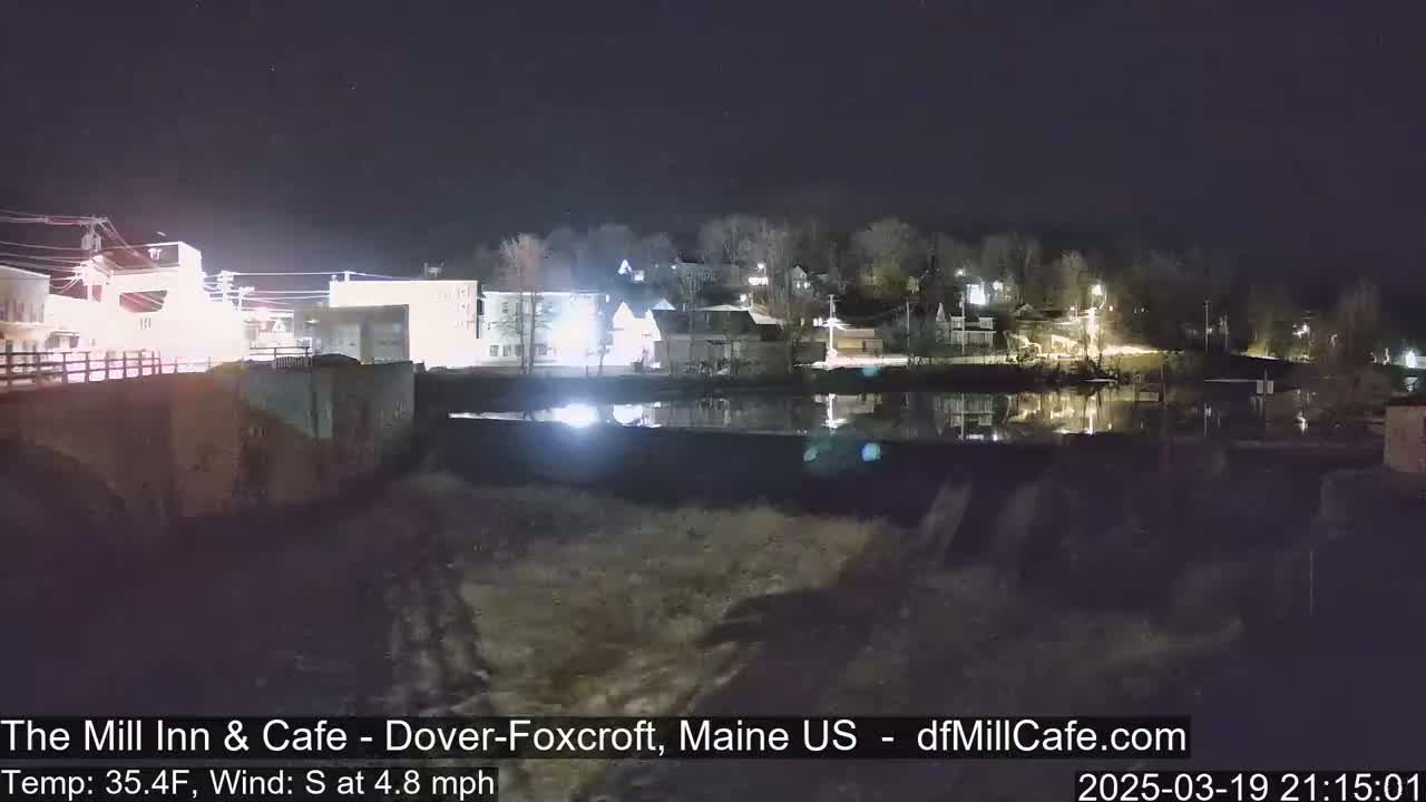A nighttime view of a calm river reflecting the lights of a small town, with a slight south wind.