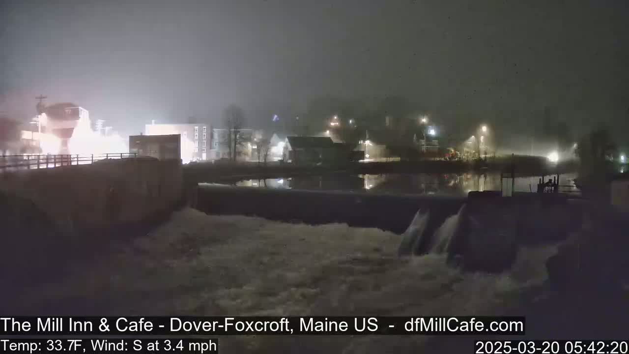 A nighttime view of a dam with flowing water under foggy conditions, showing a town's lights reflected on the water.