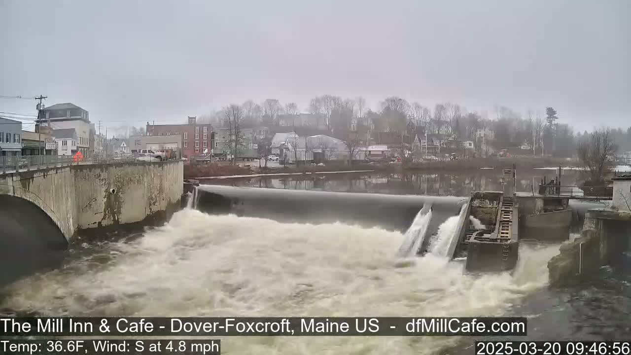 Water rushes over a dam in a town on an overcast day.