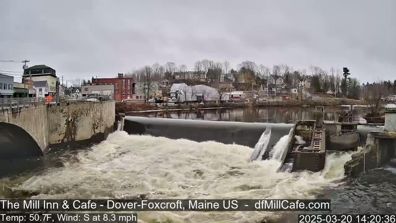 Water rushes over a dam in front of a small town under an overcast sky.