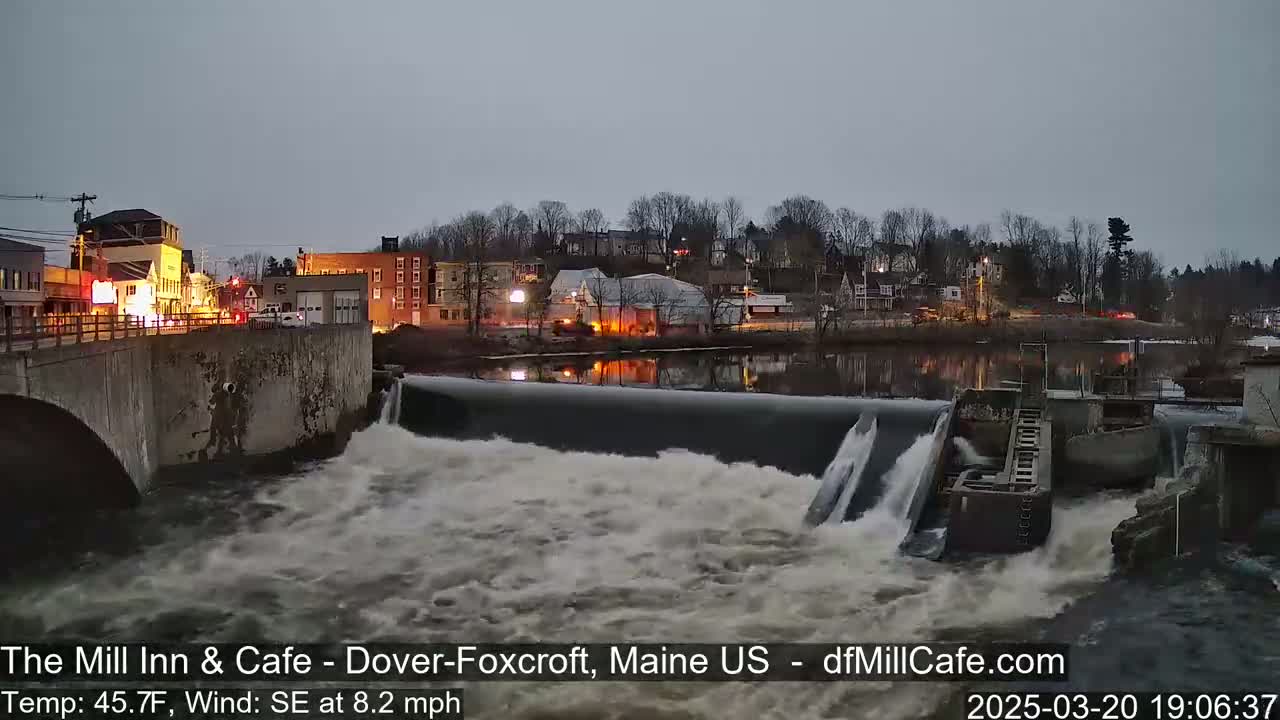 Water rushes over a dam at dusk, in front of a small town under a gray sky with a southeast wind.