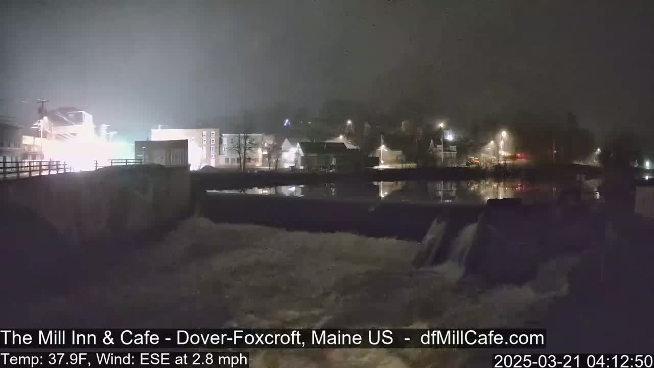 A nighttime view of a town's waterfront, showing a dam with water flowing over it under foggy conditions.