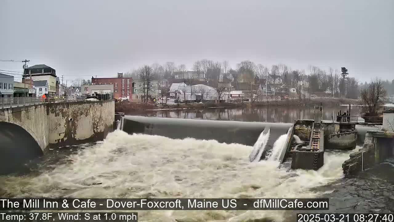 Water rushes over a dam in front of a town under an overcast sky.