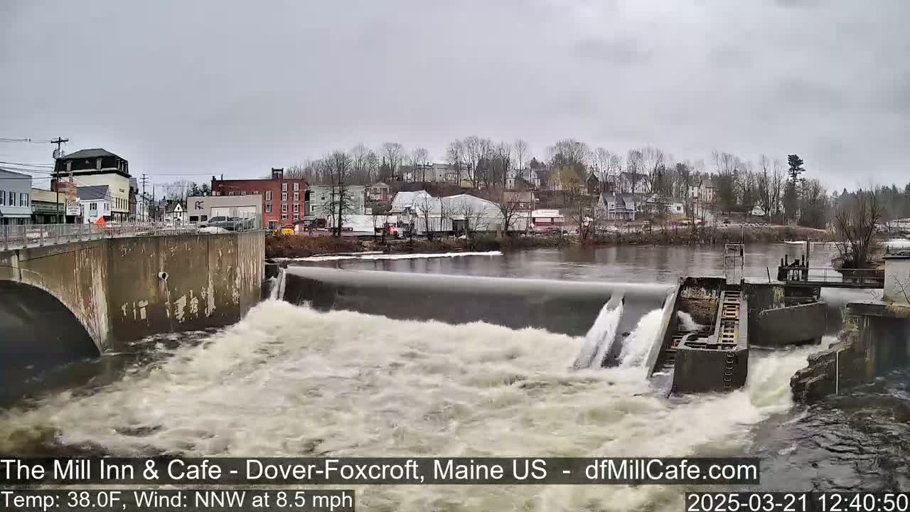 A river flows over a dam in a town on an overcast day.
