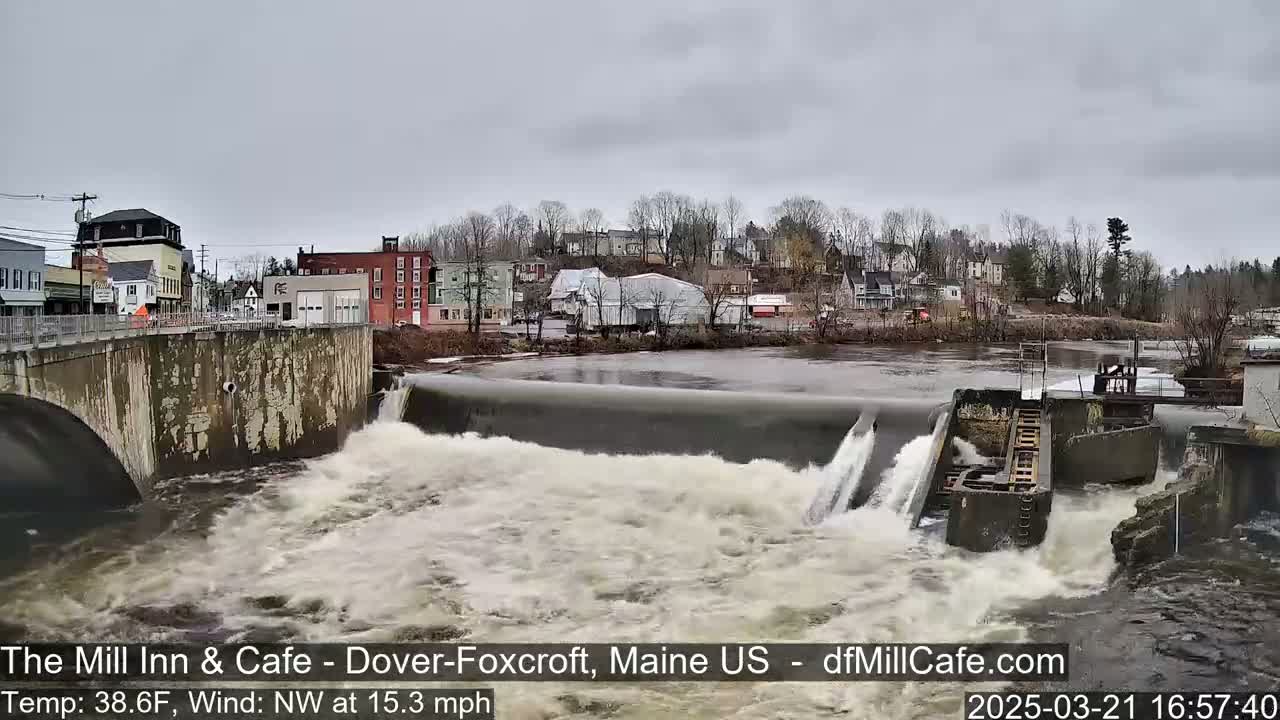A river flows over a dam in a town under an overcast sky.