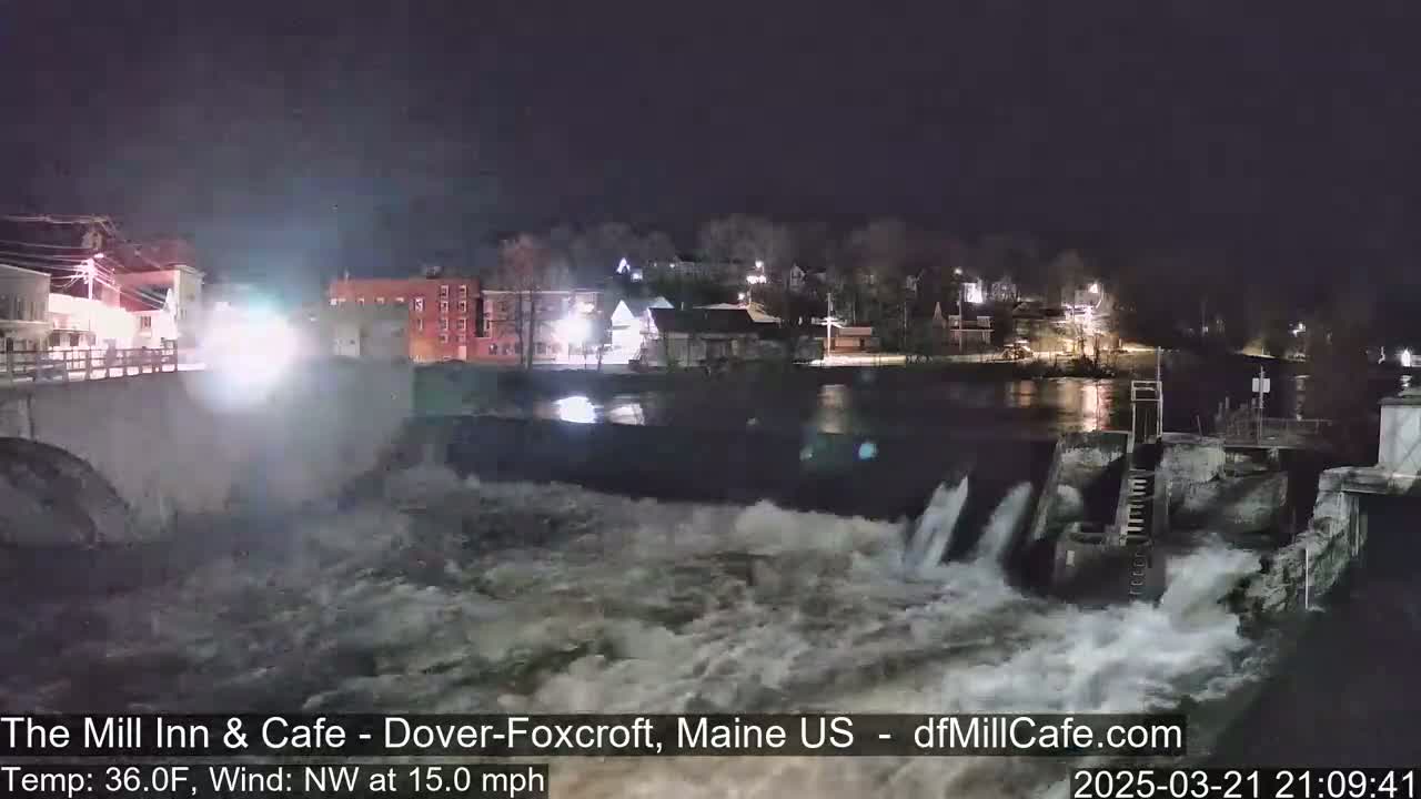 A nighttime view of a dam with rushing water flowing over it, under a dark sky with a northwest wind.