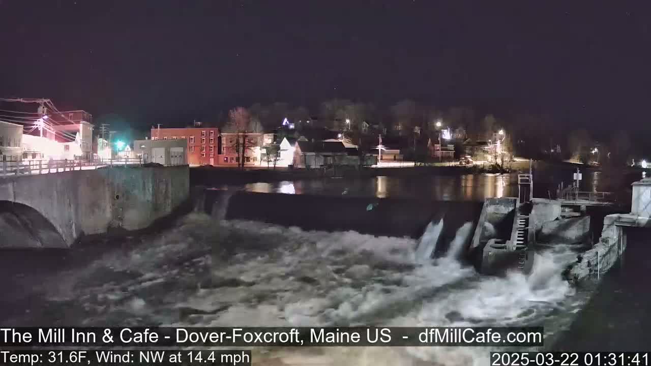 A nighttime view of a river flowing over a dam in a small town, with a northwest wind at 14.4 mph and a temperature of 31.6°F.