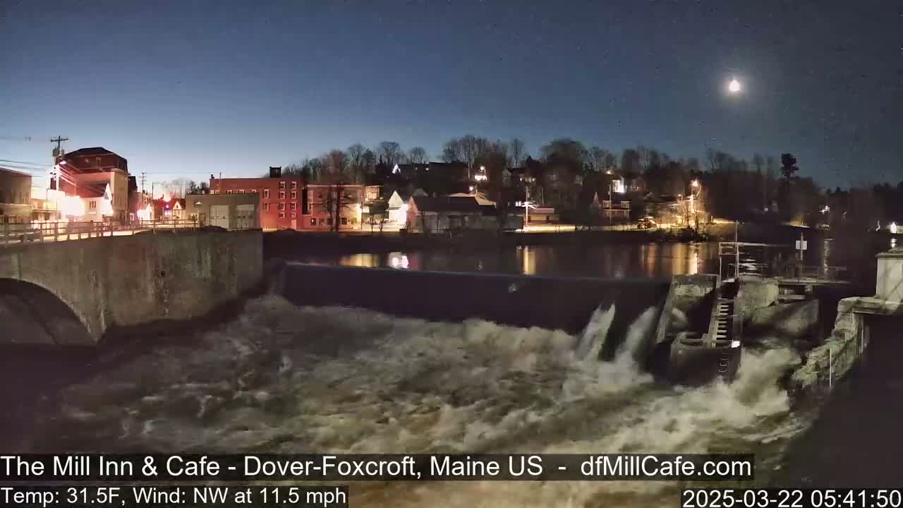 A moonlit night scene shows a dam with rushing water flowing over it, in front of a small town with lights illuminating buildings under a partly cloudy sky.