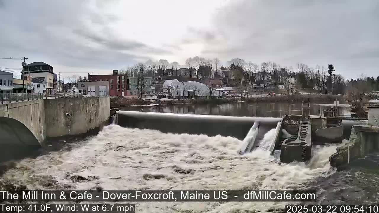 Water rushes over a dam in front of a small town under an overcast sky.