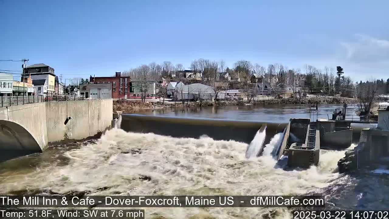 Water rushes over a dam in front of a small town on a sunny day with a southwest wind.