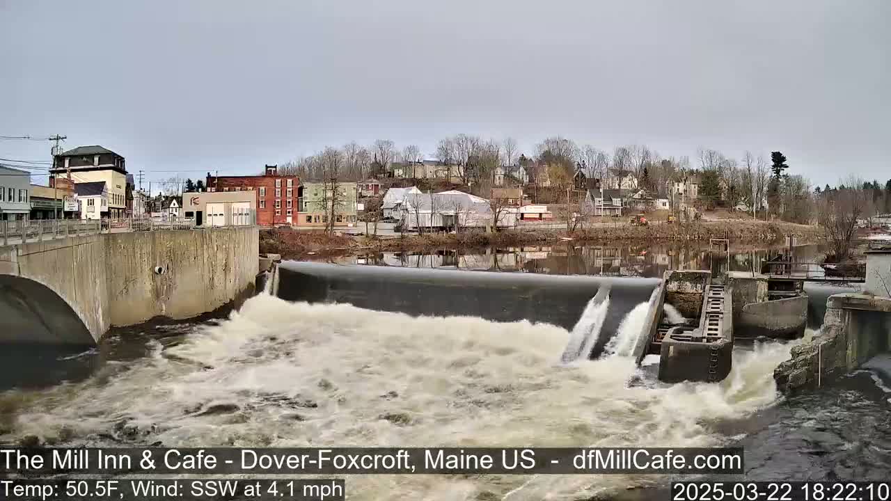 A dam with water flowing over it, in front of a town under an overcast sky.
