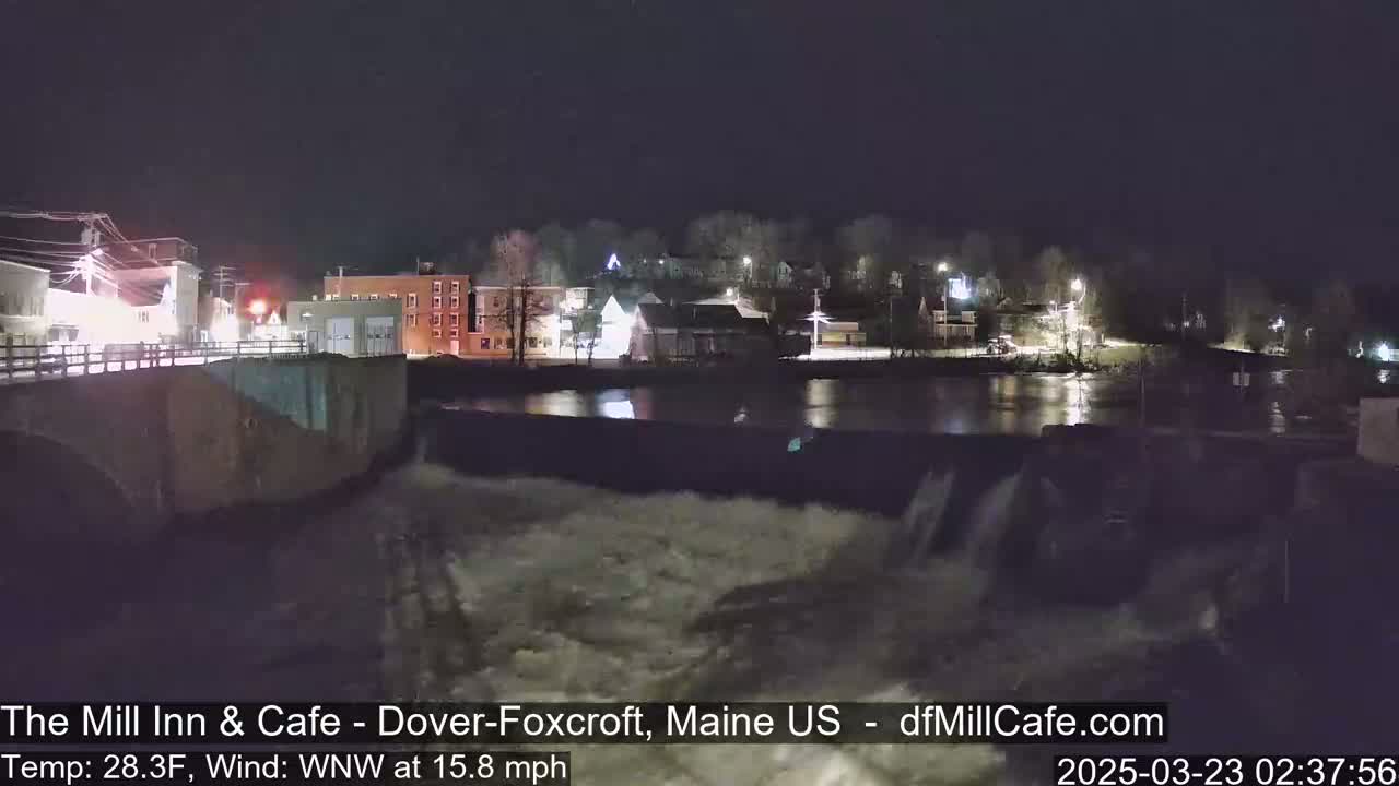A nighttime view of a river flowing over a dam in a small town, with a strong wind.
