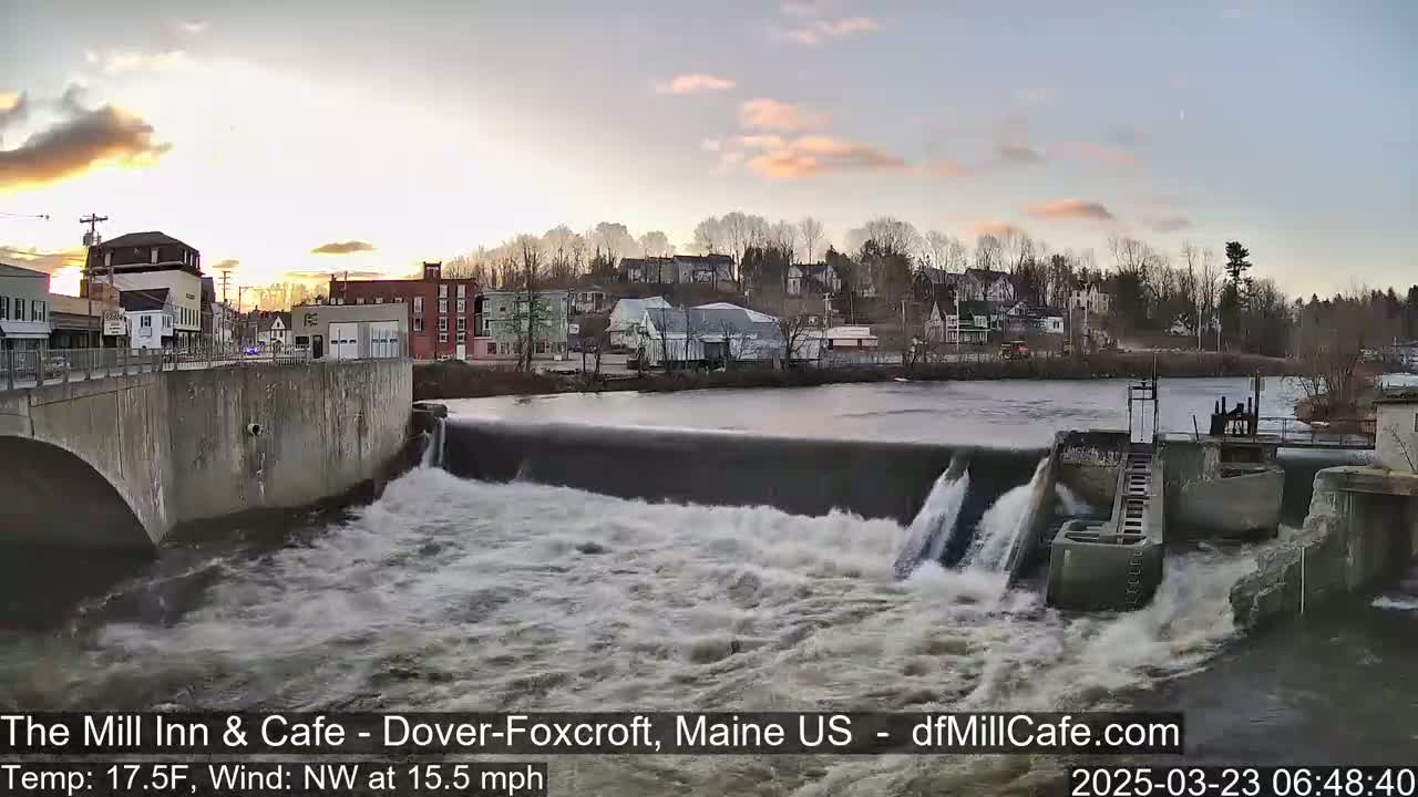 Water rushes over a dam in a town at sunset under a partly cloudy sky.