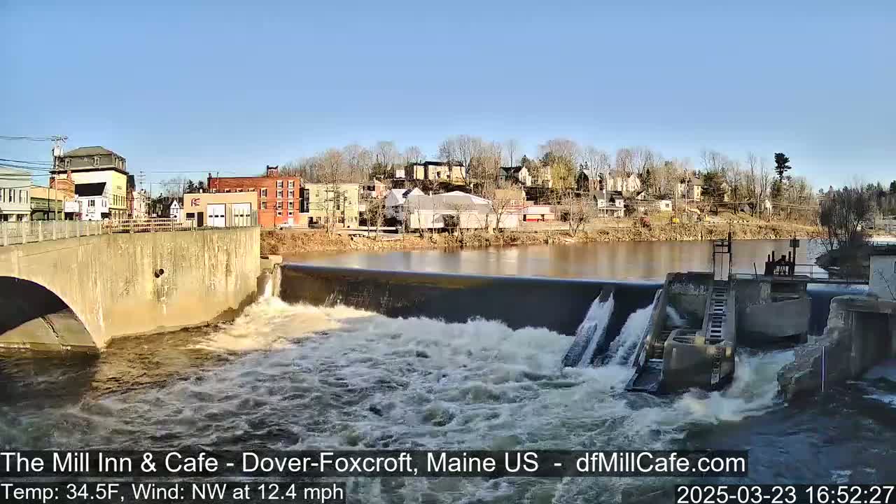 A river flows over a dam in a small town under a clear, sunny sky.