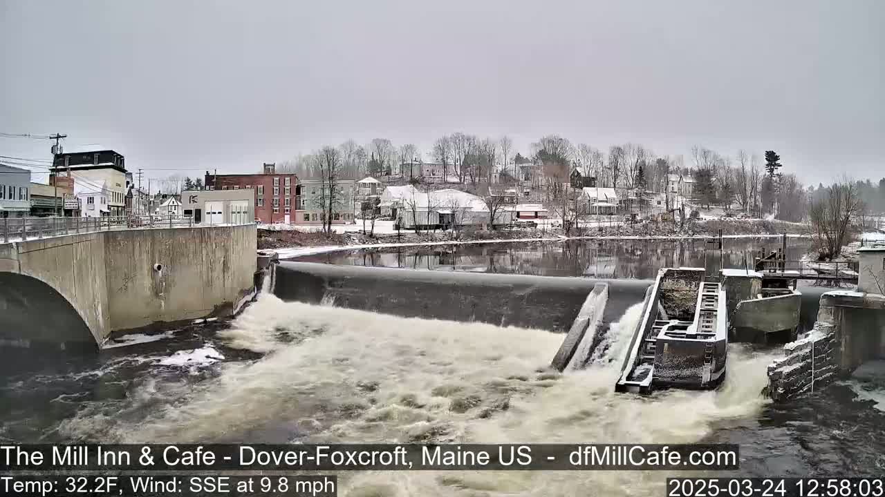 A snow-covered town is seen across a river from a dam where water is flowing over a series of gates under an overcast sky.