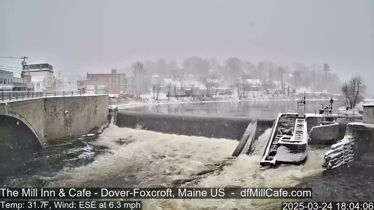 A snow-covered town is seen beyond a dam and river with a bridge in the foreground, amidst a light snowfall.