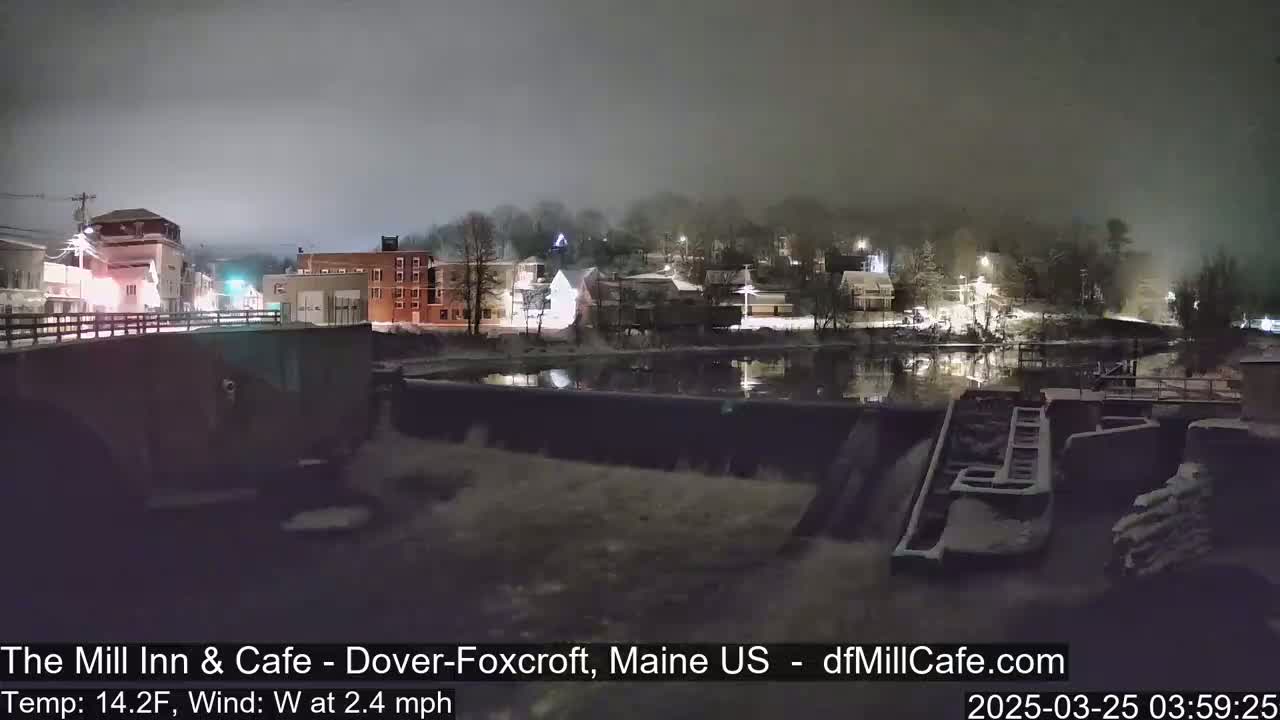A nighttime view of a snow-covered town beside a calm river and dam under a foggy sky.