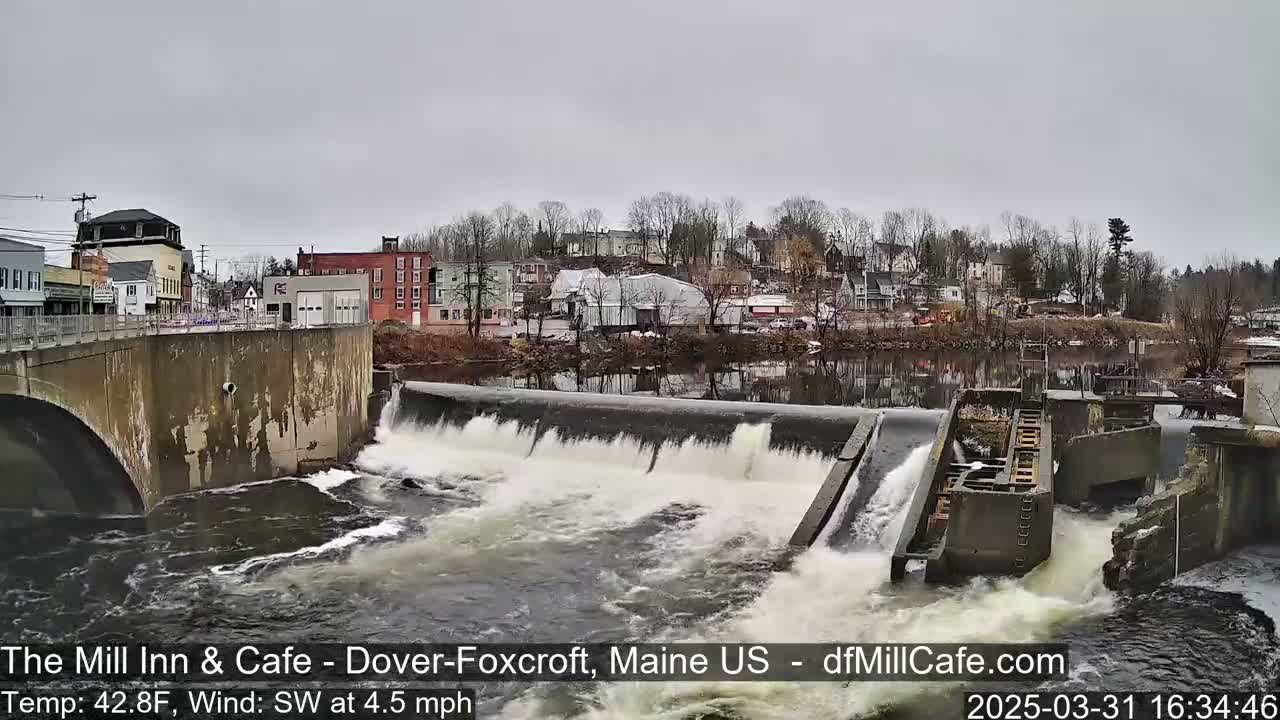 Water flows over a dam in a town under an overcast sky.