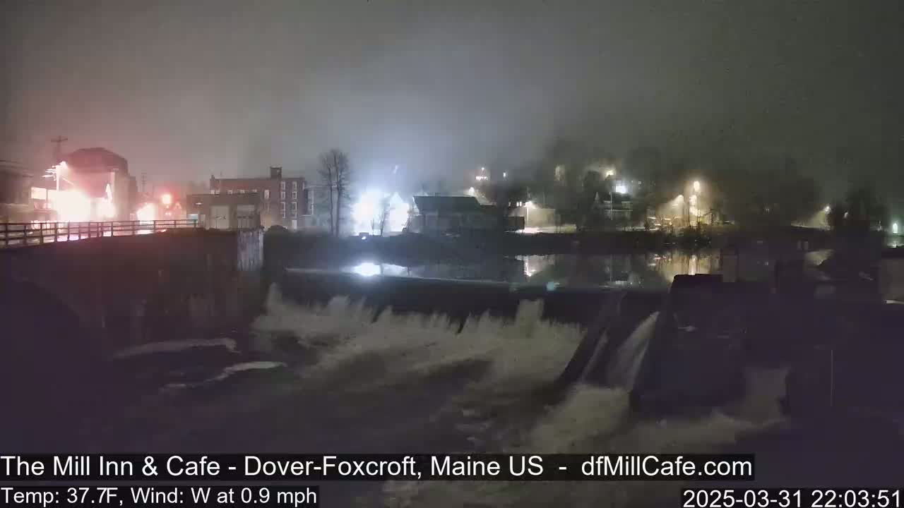 A nighttime view of a dam with water flowing over it, under foggy conditions, showing a town with lights in the background.