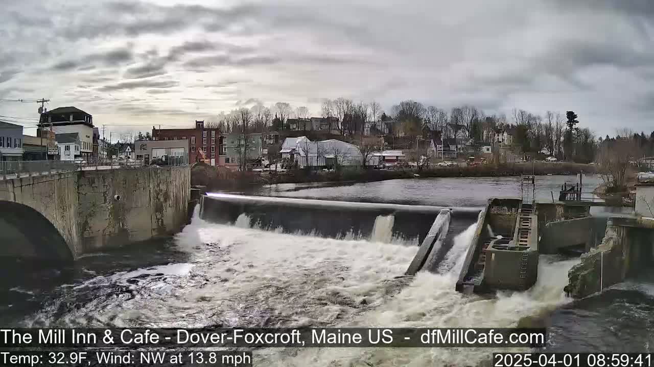 A dam with water flowing over it is situated in front of a town under an overcast sky.