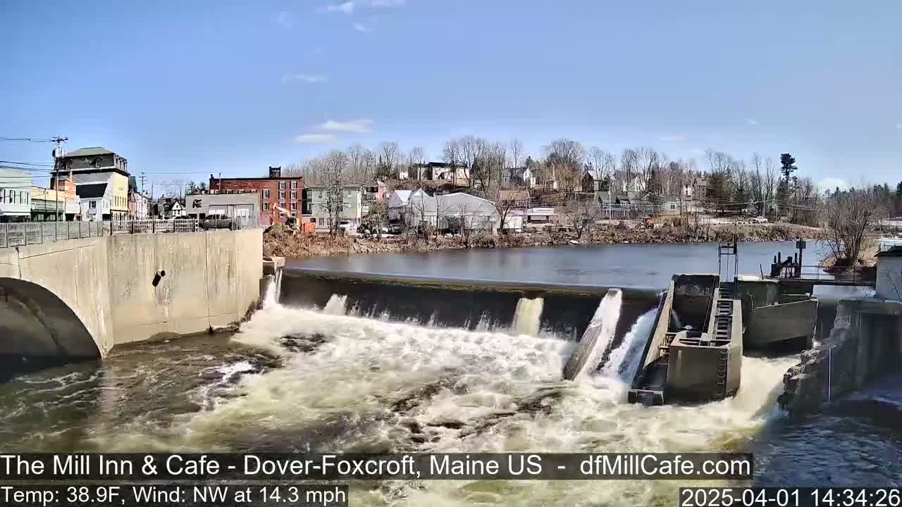 Water rushes over a dam in front of a town on a sunny day with a northwest wind.