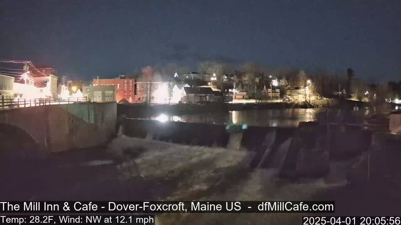 A nighttime view of a town's illuminated buildings and a dam with water flowing over it, under a clear, dark sky.