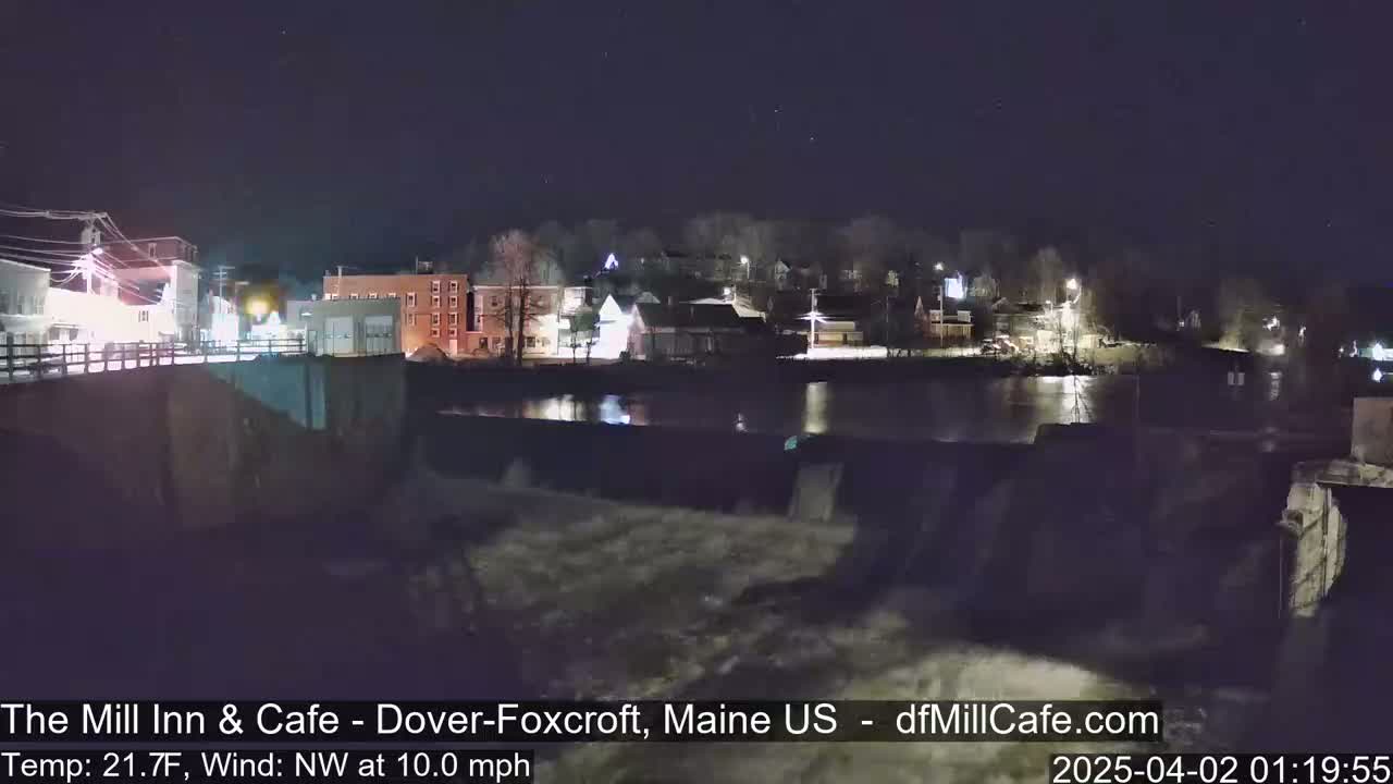 A nighttime view of a town situated next to a river with a dam, under a clear, starry sky.