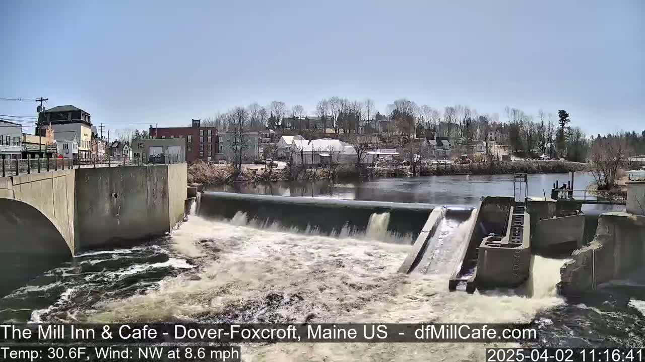 A river flows over a dam in a small town on a clear, sunny day with a northwest wind.