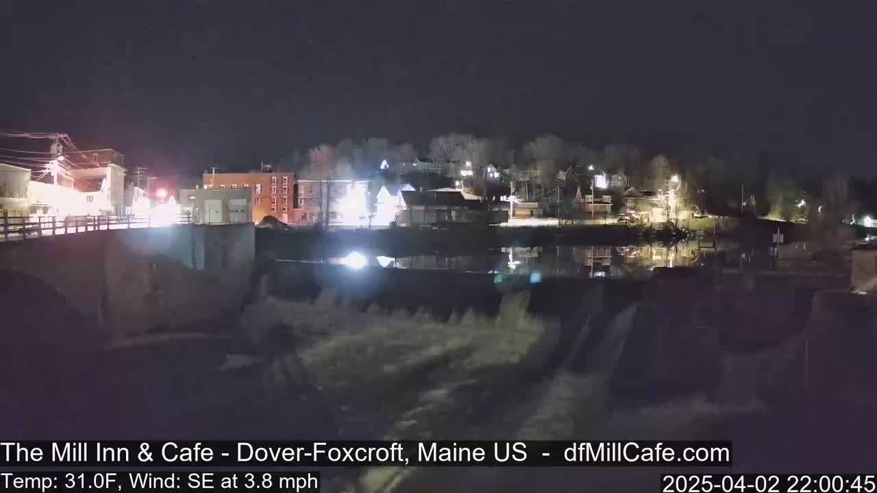A nighttime view of a town's waterfront, featuring a dam with water flowing over it, under a clear, dark sky.