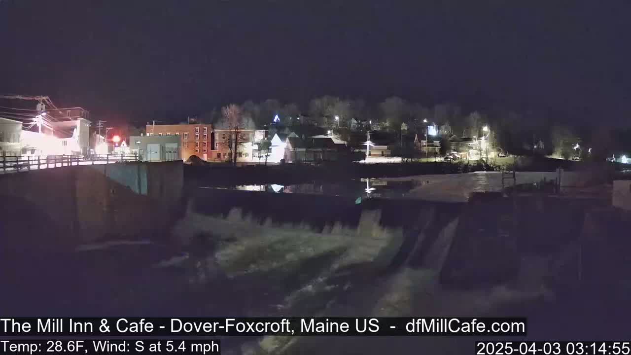 A nighttime view of a town's buildings and a dam with water flowing over it, under a clear, dark sky.