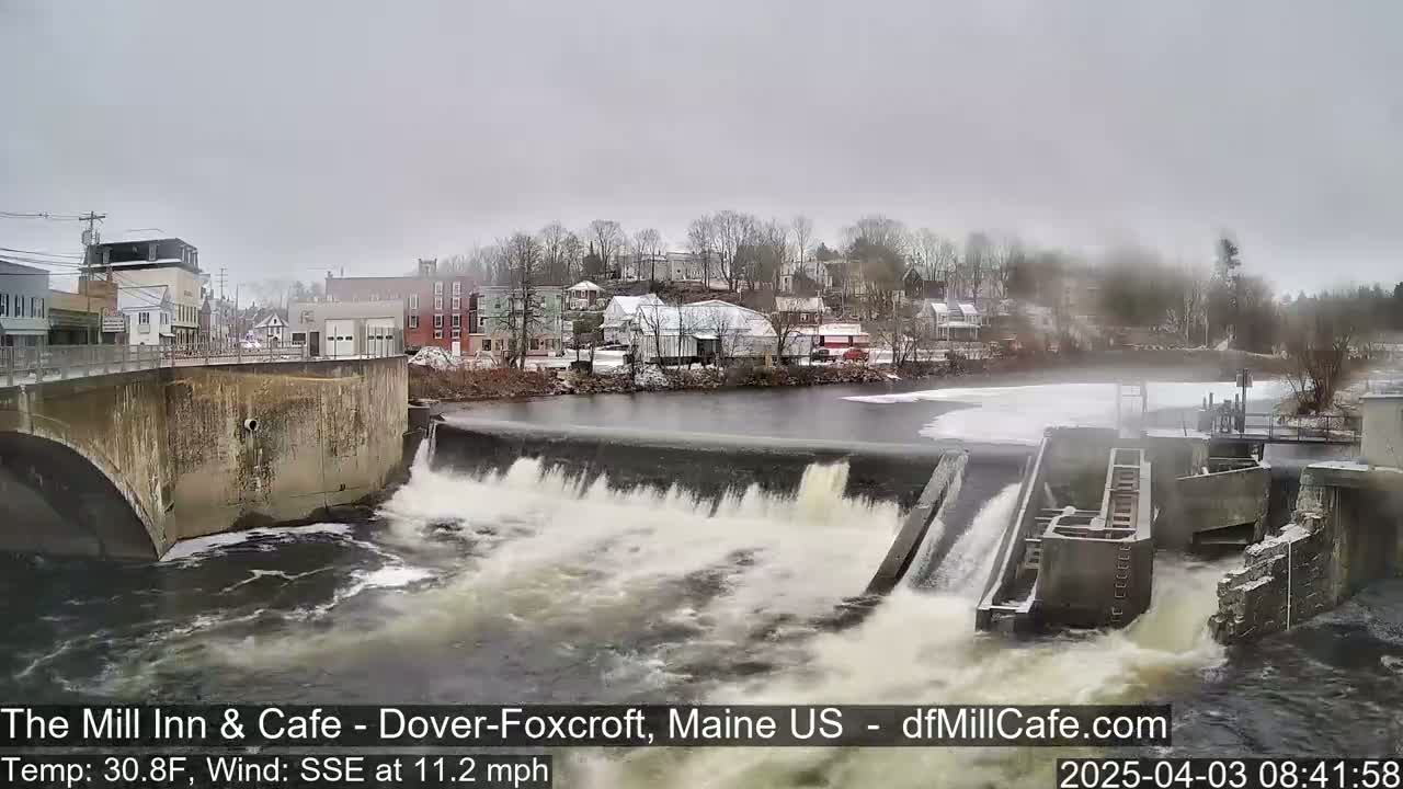 Water rushes over a dam in a town on an overcast day.