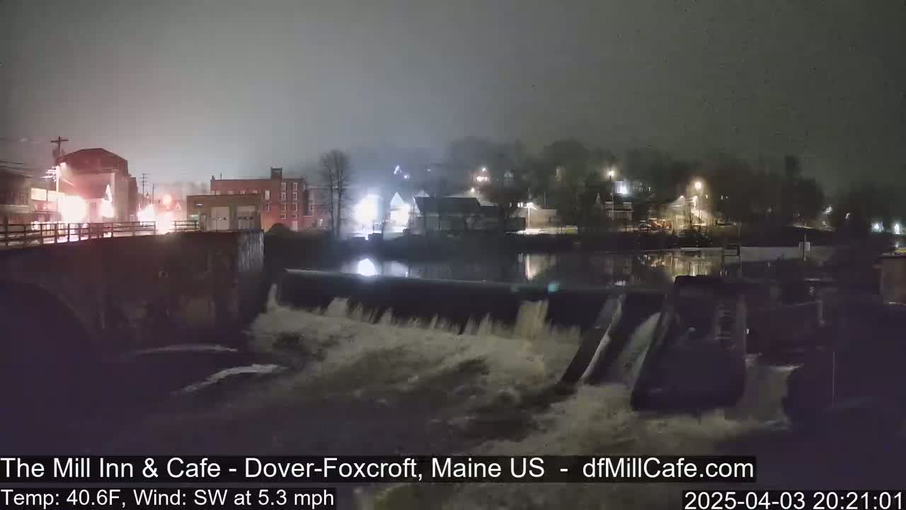 A nighttime view of a dam with water flowing over it, under foggy conditions, with lights from buildings visible in the background.