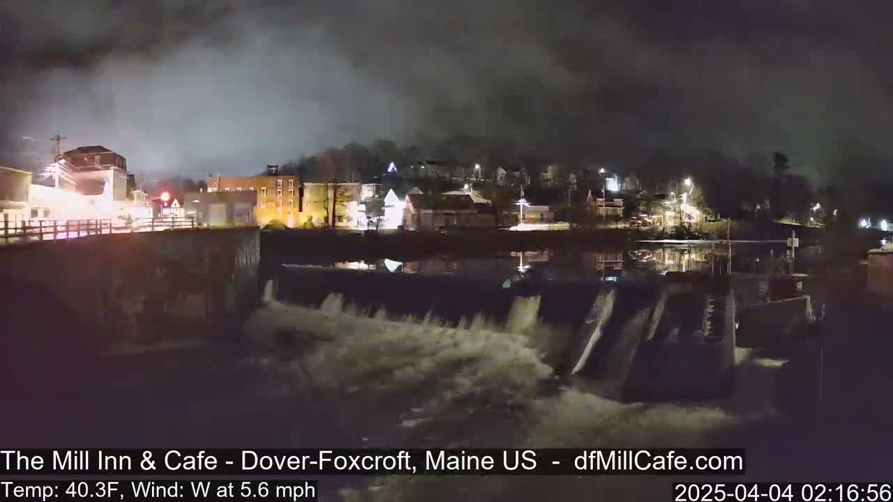 A nighttime view of a town's river dam with water flowing over it under a cloudy sky.