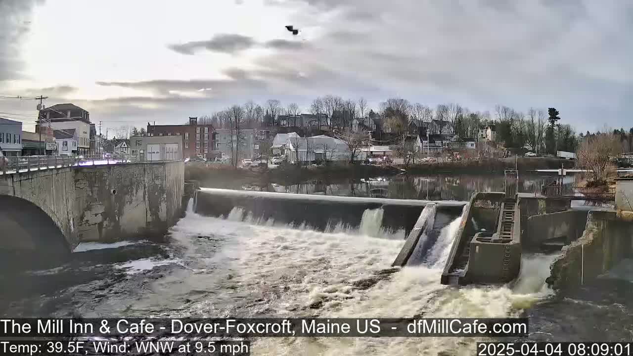Water rushes over a dam in a town under a cloudy sky.