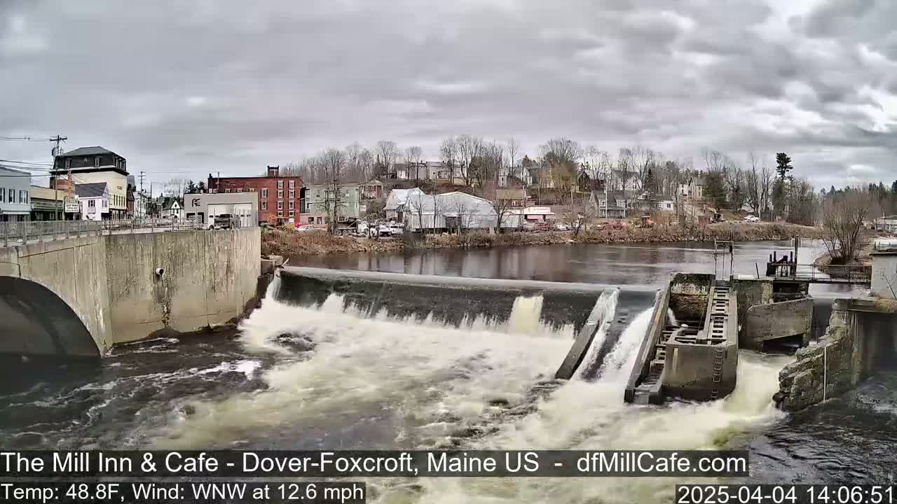 A river flows over a dam in a town under an overcast sky.
