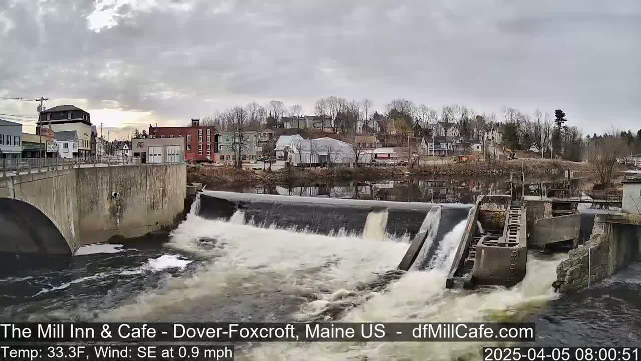 A dam releases water into a river flowing through a small town under an overcast sky.