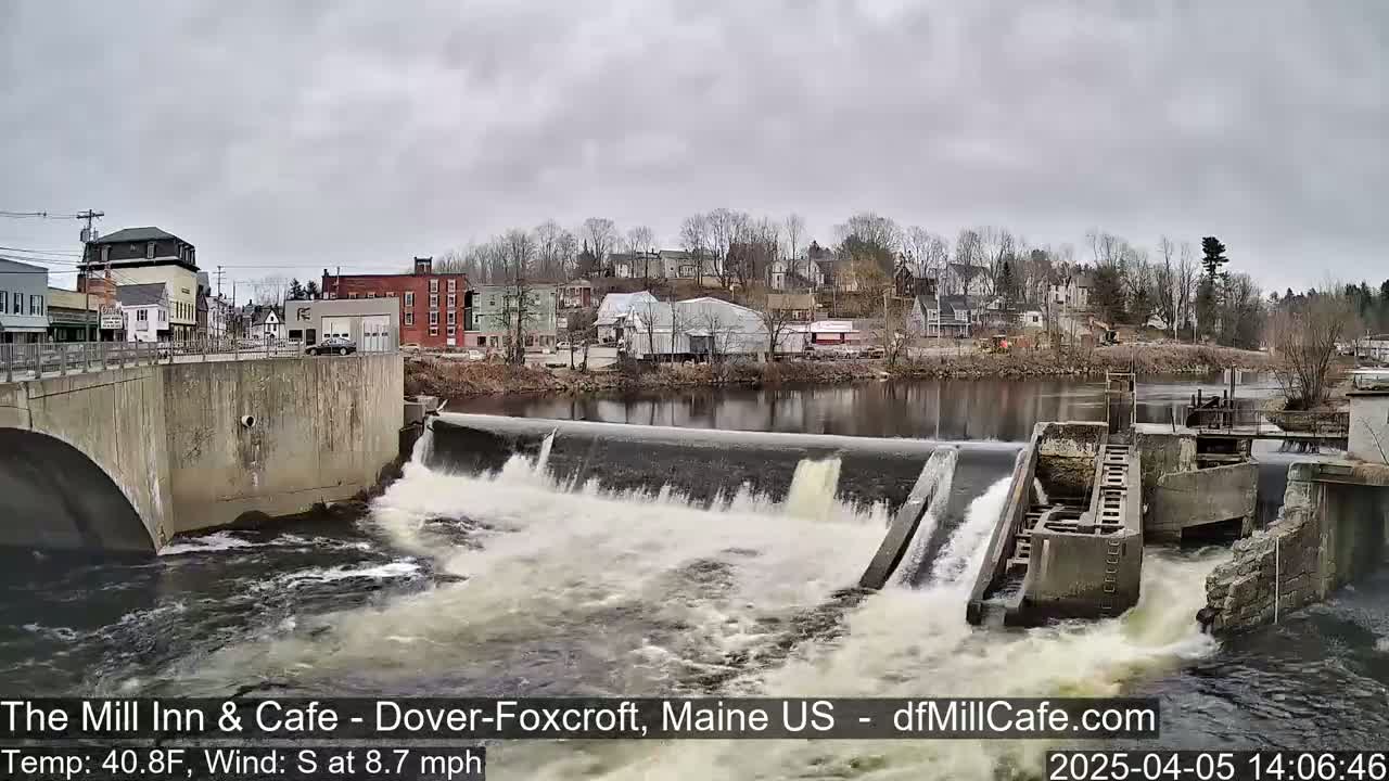 Water rushes over a dam in a town, under an overcast sky.
