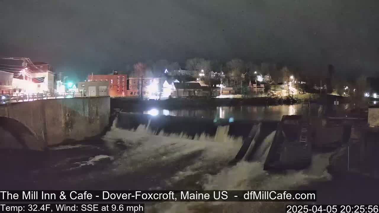 A nighttime view of a dam with water flowing over it, illuminating a riverside town under a dark sky.