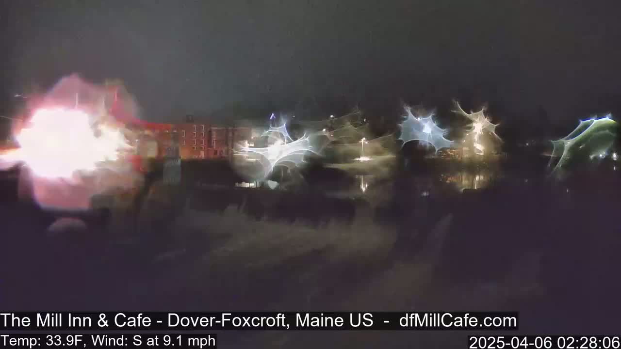 A nighttime scene shows a large bright explosion or fire in the foreground, with blurry lights and what appears to be a building in the background, under dark, possibly windy conditions.