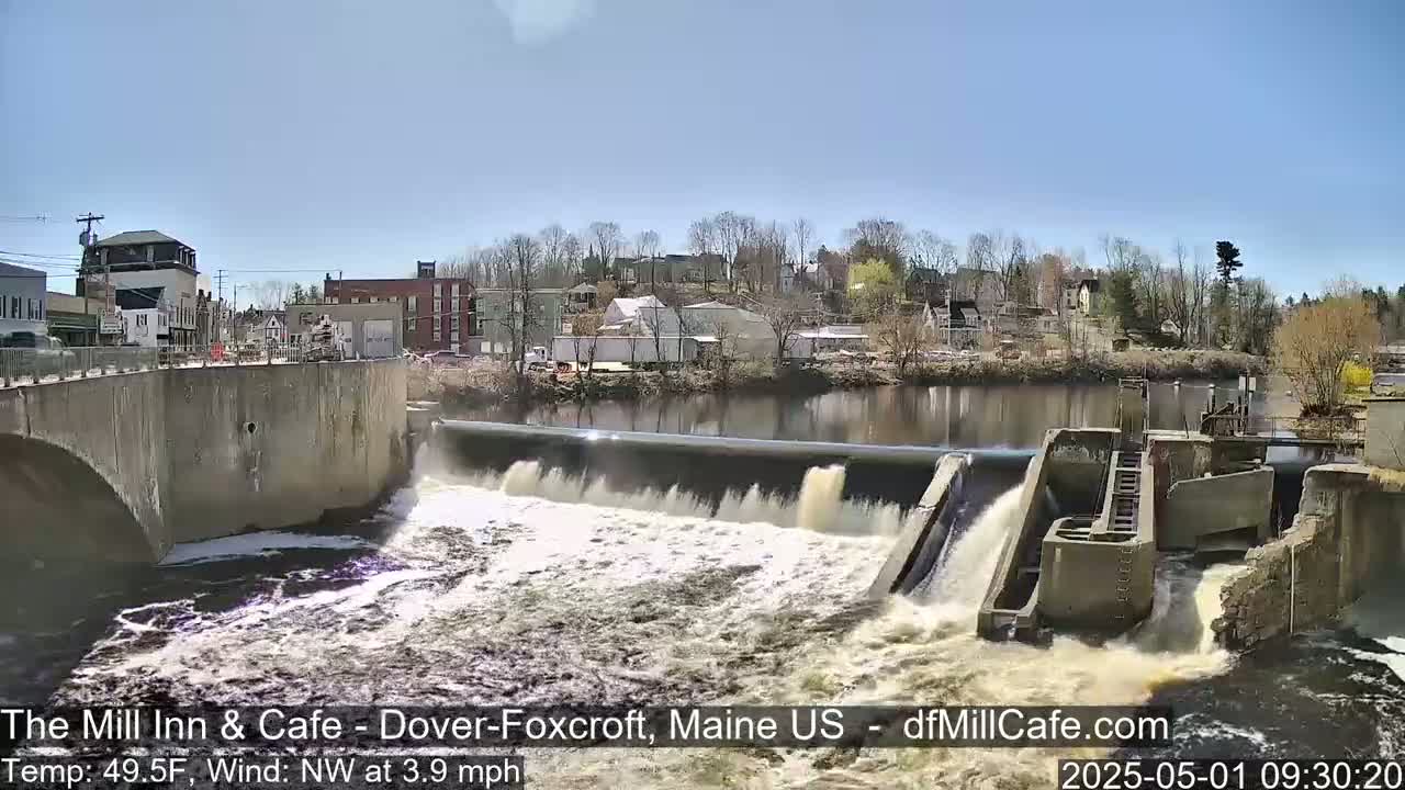 Water flows over a dam in a town on a sunny day with a light northwest wind.