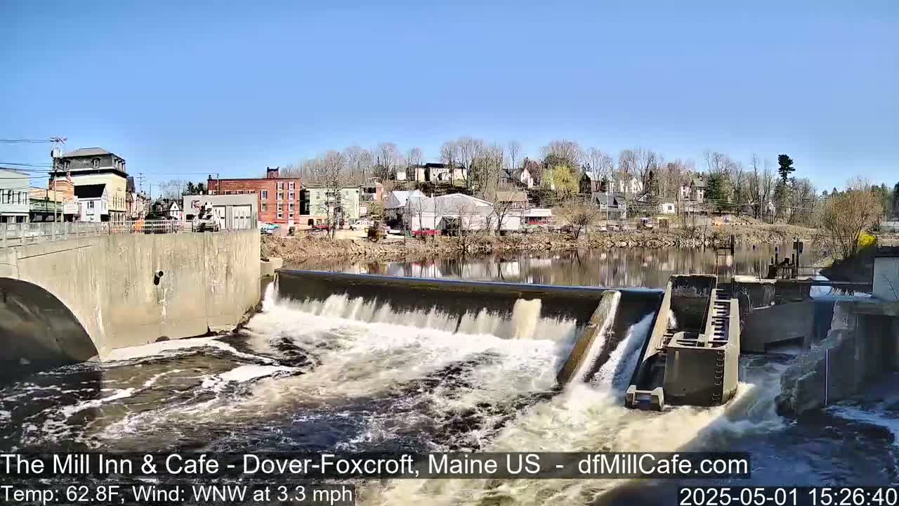 A dam releases water into a river flowing past a town under a clear, sunny sky.