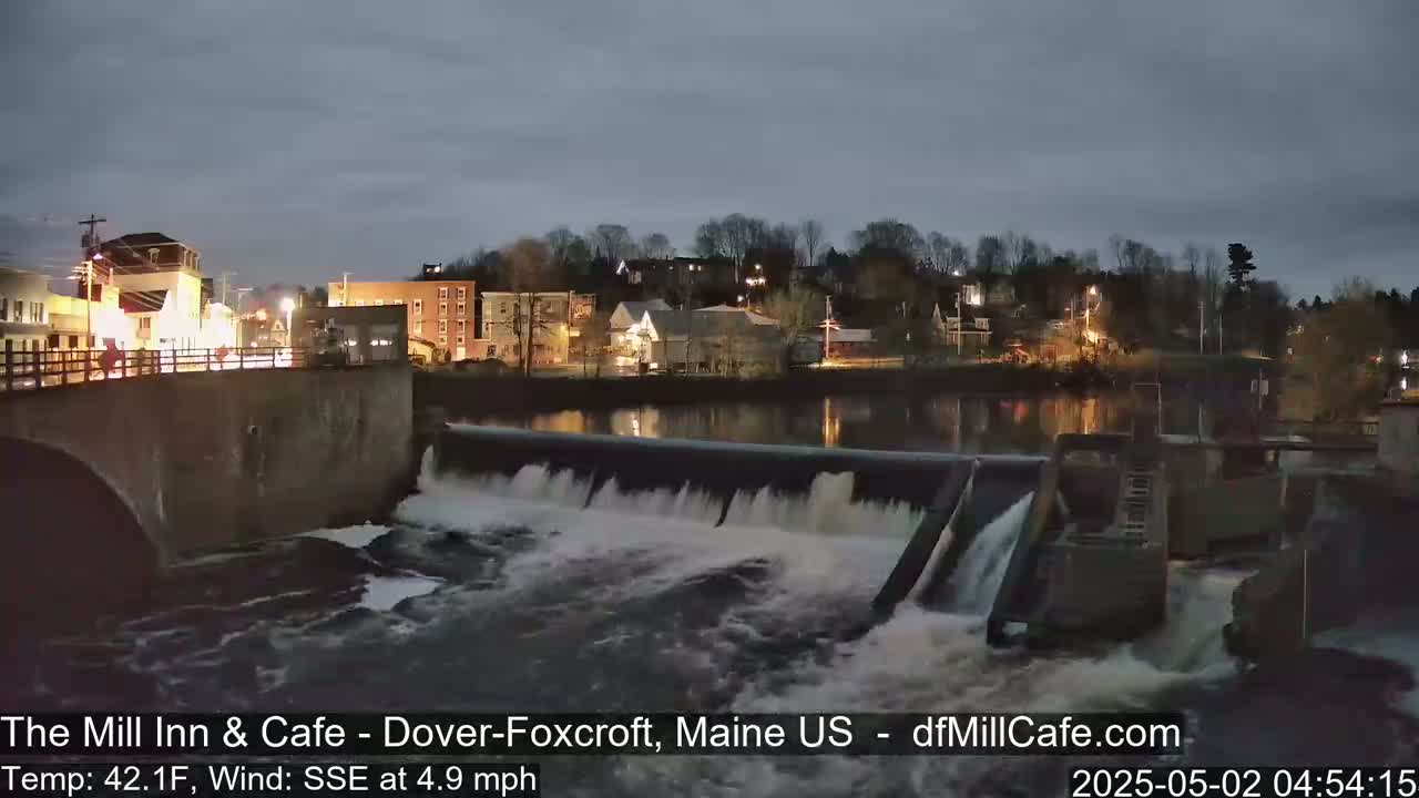 Water rushes over a dam at night, illuminating a town's lights reflected in the calm water above.